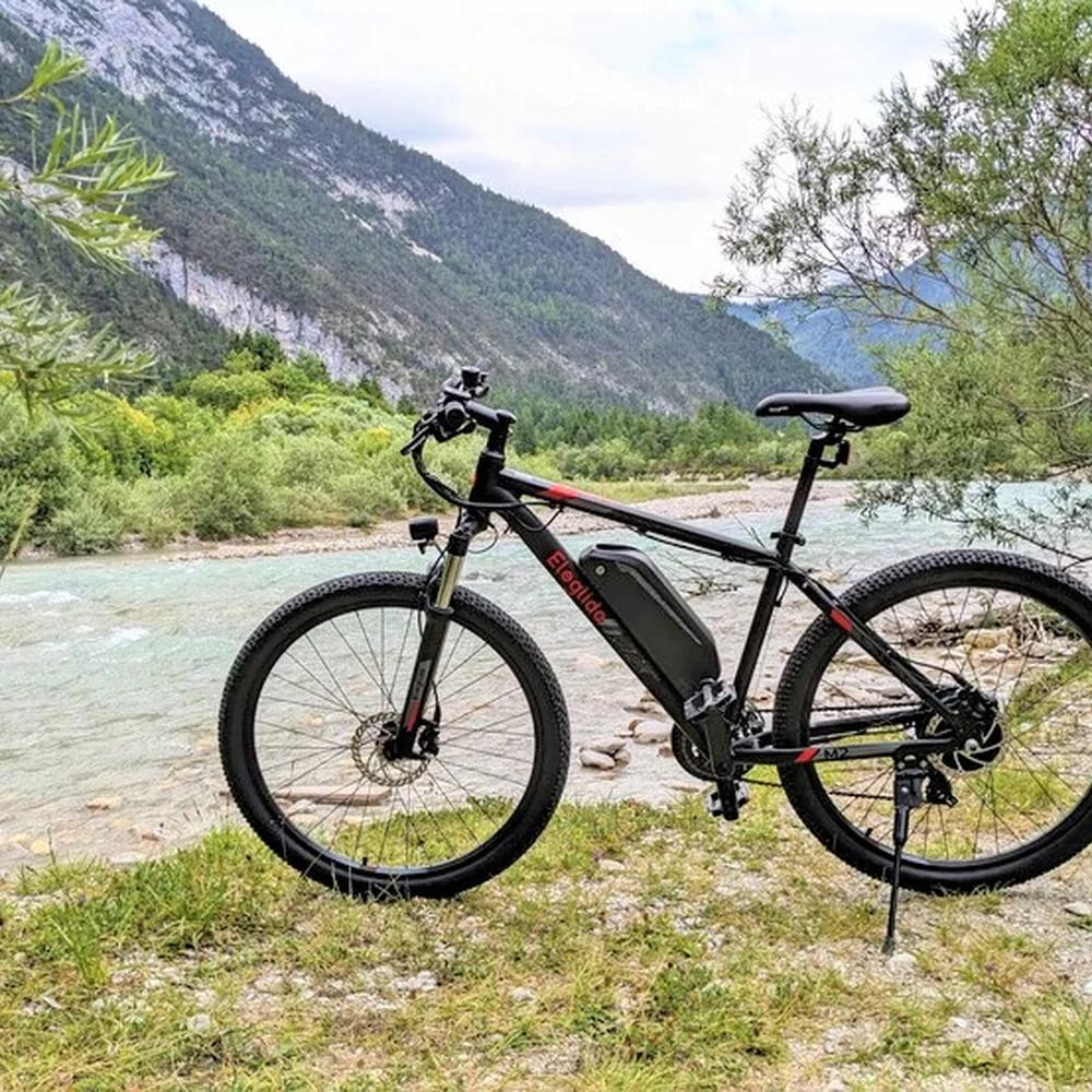 Eleglide M2 27.5" Electric Mountain Bike, 15.5MPH on its side stand next to a river in a valley in the wild countryside