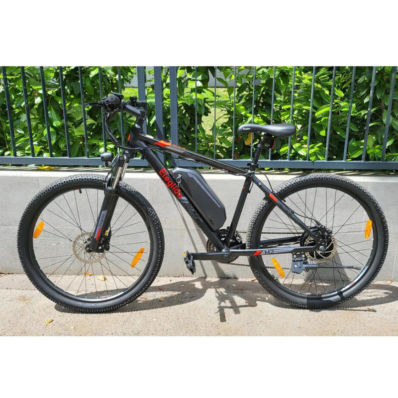 Eleglide M2 27.5" Electric Mountain Bike, 15.5MPH showing the bike on its side stand on a pavement next to a low wall and fence 